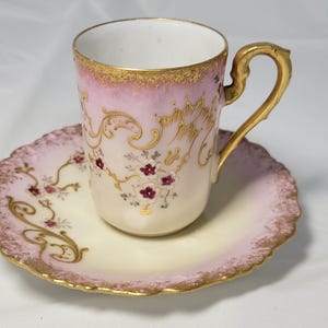 Może przedstawiać: Zestaw filiżanki ze spodkiem z porcelany o delikatnym różowym gradiencie i kremowej podstawie. Zestaw ozdobiony jest misternymi złotymi zawijasami oraz małymi skupiskami ciemnoczerwonych kwiatów. Filiżanka posiada zakrzywione złote ucho.