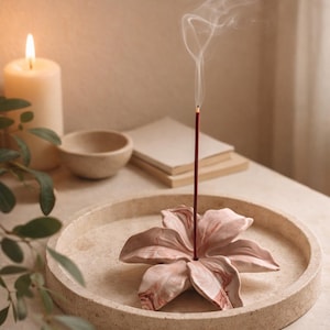May include: A circular beige stone tray containing a sculpted pink and white marbled flower incense holder. A single dark red incense stick is placed in the centre of the flower, emitting a thin trail of white smoke into the air.