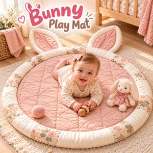 Op de afbeelding: Een ronde, gewatteerde roze speelmat met twee grote, rechtopstaande konijnenoren aan de bovenkant. De rand is versierd met een bloemenpatroon op een crèmekleurige achtergrond. De tekst Bunny Play Mat staat in roze en bruine letters boven de mat.