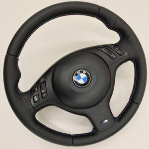 Puede incluir: Un volante BMW de cuero negro con agarres laterales perforados, costuras en contraste azul y rojo, botones de control multifunción integrados en los radios y un emblema central de BMW con una insignia M en la parte inferior.