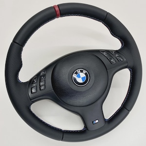 Puede incluir: Un volante BMW de cuero negro con agarres laterales perforados, una marca central de cuero rojo en la parte superior y costuras azules y rojas. El centro muestra el logotipo de BMW e incluye botones de control integrados y una insignia M.