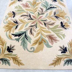 May include: A rectangular area rug with a cream-colored background featuring a large, central floral medallion and smaller corner motifs. The design uses tufted patterns in shades of sage green, mustard yellow, navy blue, and dusty pink.