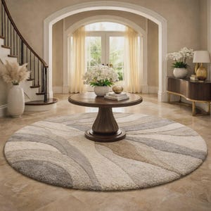 May include: A large, round area rug featuring a textured, abstract wave pattern in shades of cream, beige, and gray. The rug is positioned on a polished stone floor in a foyer, centered beneath a dark wood pedestal table with a white floral arrangement.