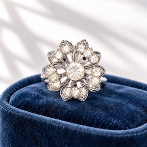 May include: A silver-toned ring featuring a central round-cut stone surrounded by a cluster of smaller round-cut stones arranged in a floral pattern. The metal setting has a polished finish, and the stones exhibit a light, clear sparkle.