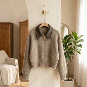 May include: A beige knit zip-up sweater featuring a contrasting dark brown leather-look collar and a gold-toned metal zipper. The garment has long sleeves with ribbed cuffs and a matching ribbed hem, providing a structured and clean silhouette.