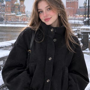 May include: A black jacket with a high collar, long sleeves, and a front button closure. The jacket features a cinched elastic hem at the waist and is worn with a black shoulder bag with a gold buckle, set against a snowy outdoor backdrop.
