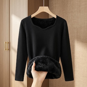 May include: A long-sleeved black shirt with a square neckline, featuring a thick, plush, black faux fur lining visible at the hem. The garment is displayed on a wooden hanger, showcasing the soft, insulating interior texture of the fabric.