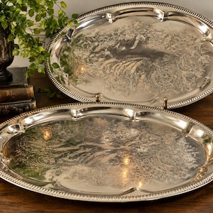 May include: Two oval silver-toned serving trays featuring a scalloped edge with a beaded border. The center of each tray displays an intricate, etched floral and scrollwork pattern across the polished metallic surface.