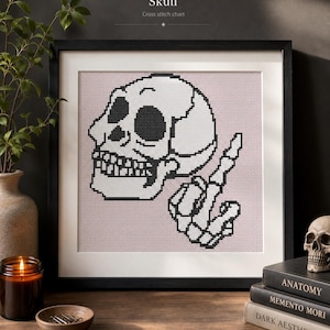 Könnte beinhalten: Ein gerahmtes Kreuzstichmuster mit dem Titel Skull, das eine schwarz-weiße, verpixelte Illustration eines menschlichen Schädels und einer skelettierten Hand mit einer Geste vor hellrosa Hintergrund in einem schwarzen Rahmen zeigt.