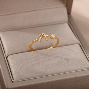 Puede incluir: Un anillo abierto de tono dorado que presenta una letra A mayúscula en un extremo y una pequeña piedra preciosa transparente de corte redondo engastada en el otro extremo. La banda es fina y circular, con un espacio ajustable entre ambos.
