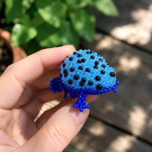 May include: A small, three-dimensional frog figurine crafted from tiny beads. The frog features a bright blue, rounded back covered in small, evenly spaced black beads, while the legs and underside are made of a solid, deep royal blue beadwork.
