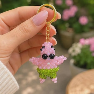 Könnte beinhalten: Ein Perlen-Schlüsselanhänger in Form einer rosa Seestern-Figur mit schwarzen Augen und grüner Hose. Die Figur besteht aus kleinen, durchscheinenden rosa und grünen Perlen und ist mit einer kurzen goldfarbenen Kette an einem goldenen Schlüsselring befestigt.
