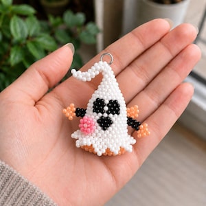 Può includere: Un ciondolo a forma di fantasma realizzato con piccole perline bianche, nere, arancioni e rosa. Il fantasma presenta occhi neri a forma di cuore, un piccolo accento circolare rosa e dettagli in perline arancioni sulle braccia e sulla base, con un anello in metallo argentato.