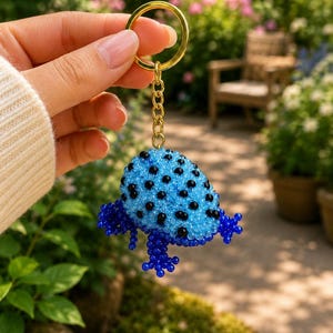 Peut inclure: Un porte-clés en perles représentant une figurine de grenouille en trois dimensions, fabriquée à partir de petites perles bleu vif et noires. La grenouille a un dos bleu clair avec des points noirs et des pattes bleu foncé, fixé à un anneau doré.