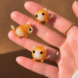 May include: Three small, round cat figurines crafted from tiny orange and white beads. Each cat features black bead eyes, a small pink bead nose, pointed ears, and a short tail. The figures are spherical in shape with a textured, beaded surface.