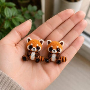 Puede incluir: Dos pequeñas figuras de panda rojo hechas a mano con diminutas cuentas uniformes. Cada figura presenta una cabeza de color naranja brillante, marcas faciales blancas, grandes ojos negros y un cuerpo marrón oscuro con una cola a rayas.