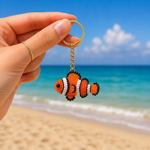 May include: A beaded clownfish keyring featuring vibrant orange, white, and black glass beads arranged in a detailed fish pattern. The charm is attached to a gold-toned metal split ring by a short matching chain, creating a small decorative accessory.