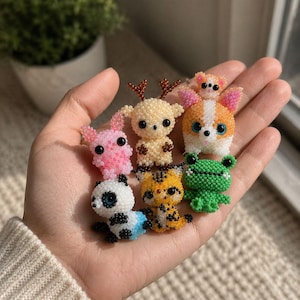 Puede incluir: Una colección de siete figuras de animales en miniatura hechas con pequeñas cuentas de colores. El conjunto incluye un conejo rosa, un ciervo beige con astas marrones, un perro blanco y naranja, un panda, un tigre, una rana verde y una criatura blanca.