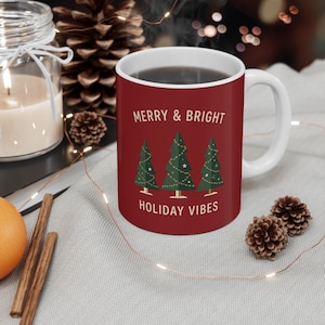 Op de afbeelding: Een donkerrode keramische mok met een witte binnenkant en handvat. De voorzijde toont drie groene kerstbomen versierd met gouden lichtjes en sterren, met de tekst MERRY & BRIGHT erboven en HOLIDAY VIBES in het wit eronder gedrukt.