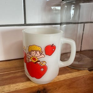 May include: White ceramic mug featuring a cartoon character with yellow hair and a yellow outfit, riding a red apple. The mug has a handle and a vintage design. A clear glass jar with a metal lid is in the background.
