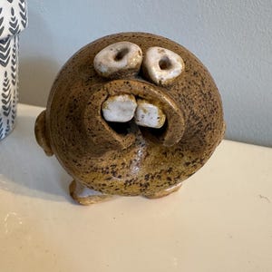 May include: A whimsical, handmade ceramic sculpture of a round, brown-speckled face with large, off-white eyes and two prominent teeth. The sculpture has small ears and a wide, expressive mouth, creating a humorous and unique decorative piece.