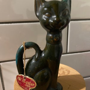 May include: A vintage ceramic cat figurine with a dark green and teal glaze. The cat has a smiling expression and a handle on its back. A tag reads "Mc Master craft".