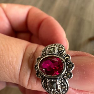 May include: A silver-toned ring featuring an oval, deep red gemstone as the centerpiece. The ring is adorned with smaller, round, and square accents, possibly marcasite stones, set around the main stone and on the band.