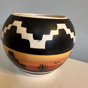May include: A round, hand-painted ceramic bowl with a black upper section featuring a white geometric pattern. The lower portion displays a sunset scene with orange, yellow, and brown hues, and a stylized tree and bird.