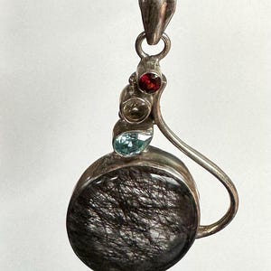 May include: Silver pendant with a large, round, black stone with hair-like inclusions. The pendant features three smaller gemstones: red, yellow, and blue. The pendant hangs from a silver chain.