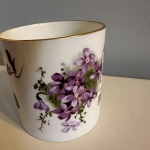May include: White ceramic cup with a gold rim, adorned with a cluster of purple flowers and green leaves. The floral design is painted on the side of the cup, creating a vintage aesthetic. The cup is likely for drinking.