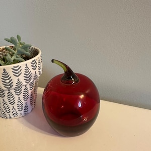 May include: A decorative glass apple with a deep red body and a dark brown base. The apple has a green stem and sits on a white surface. A patterned white and gray pot with a succulent plant is to the left.
