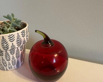 May include: A decorative glass apple with a deep red body and a dark brown base. The apple has a green stem and sits on a white surface. A patterned white and gray pot with a succulent plant is to the left.
