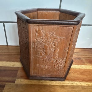 May include: A wooden, hexagonal container with a dark brown rim and base. The container features a carved landscape scene with mountains, trees, and figures. The wood has a light brown color with darker brown accents.