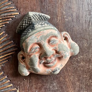 May include: A weathered, smiling face sculpture with a hat. The face is a mix of peach, gray, and black, with visible signs of age and wear. The hat has a textured pattern. The sculpture is likely made of stone or a similar material.