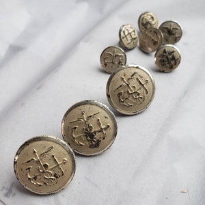 May include: A collection of nine silver-toned buttons with a raised design of an anchor, wings, and a cross. The buttons are arranged in a row on a white surface.