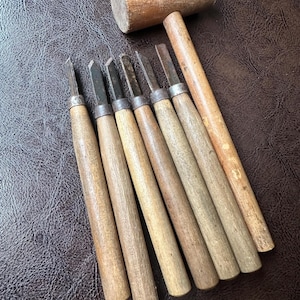 May include: A set of six wood carving chisels with light brown wooden handles and metal ferrules, accompanied by a cylindrical wooden mallet with a long handle. The tools are arranged in a parallel row, showcasing their natural wood grain.