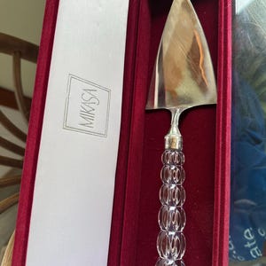 May include: A silver cake server with a clear, decorative handle, presented in a red velvet box. The handle features a series of clear, rounded segments. The box has a white satin lining and the "MIKASA" logo.