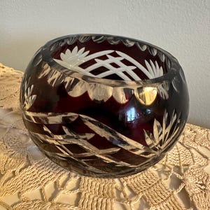 May include: A round, ruby-colored crystal bowl with intricate cut designs. The bowl features a clear, decorative pattern around the rim and etched leaf and geometric patterns on the sides. The bowl sits on a cream-colored, crocheted surface.