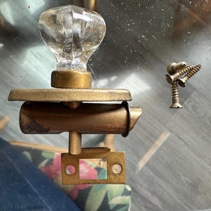 May include: Antique door hardware set featuring a clear glass knob with a brass base, a brass latch, and a separate decorative screw assembly. The hardware is displayed on a glass surface.