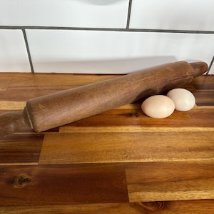 May include: A cylindrical wooden rolling pin with a smooth, dark brown finish and rounded handles at both ends, positioned horizontally next to two light beige, oval-shaped eggs. The rolling pin shows a natural wood grain texture throughout.