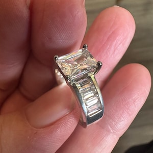 May include: A silver ring featuring a large, square-cut, clear gemstone as the centerpiece. The band is adorned with smaller, rectangular-cut stones. The ring is held in a hand, showcasing its design and sparkle.