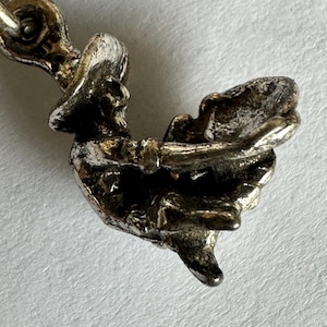 May include: Close-up of a small, detailed silver charm in the shape of a bird. The charm has intricate detailing, with visible wings and a beak. The metal has a slightly tarnished appearance, suggesting age or a vintage style.