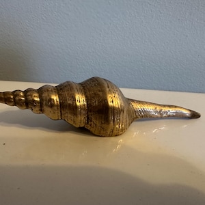 May include: A golden-colored, decorative seashell sculpture. The shell has a spiral shape with a textured surface and a pointed end. The sculpture is resting on a white surface.