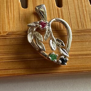 May include: A silver pendant in the shape of a heart with a floral design. The pendant features a red, green, and black gemstone. The pendant is on a wooden surface.