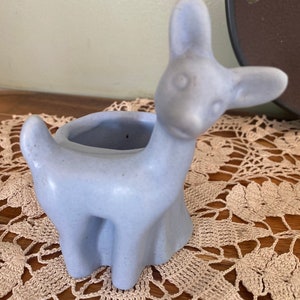 May include: A light blue ceramic deer planter with a small opening for planting. The deer is standing on a crocheted doily.