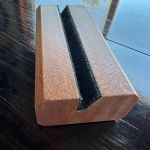 May include: A wooden phone stand with a dark brown finish. The stand has a rectangular shape with a groove lined with black felt. The stand is designed to hold a phone or tablet.