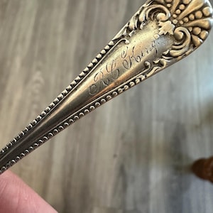May include: A silver-toned metal spoon handle featuring an ornate, embossed fan-like pattern at the top and a beaded border along the edges. The handle is engraved with the cursive text R.C. King in the center of the smooth, polished surface area.