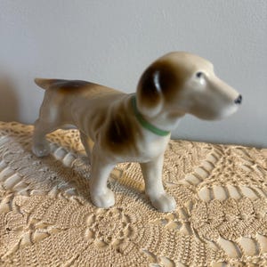 May include: A ceramic dog figurine in a standing pose. The dog has a tan and brown coat with a green collar. The figurine is on a cream-colored, crocheted surface. The dog's features are detailed, with a small black nose.