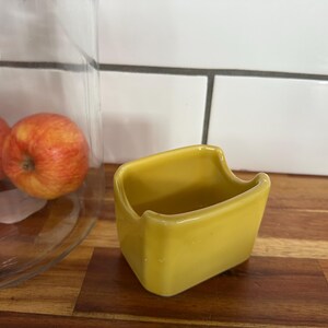 May include: A small, yellow ceramic container with a curved rim. The container is sitting on a wooden surface. In the background, there are red apples and a glass dome.