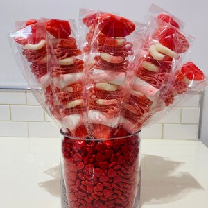 Op de afbeelding: Een glazen vaas gevuld met rode hartvormige snoepjes. Vijf heldere, in plastic verpakte snoepspiesjes zijn in de vaas gerangschikt. Elke spies is bedekt met een rood snoepje in de vorm van een lippenpaar. De spiesjes zijn gevuld met rood en wit snoep.